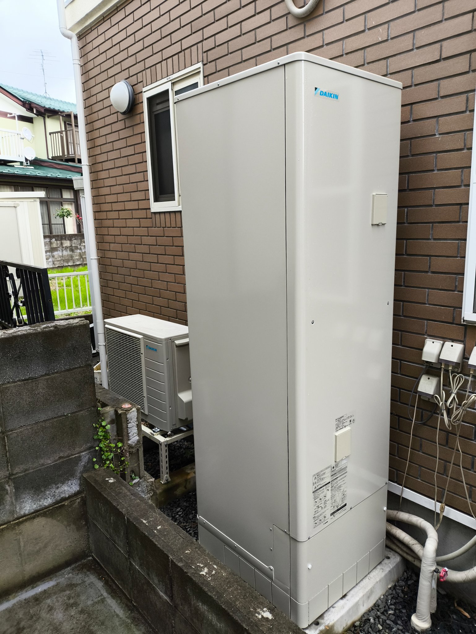 住宅の外壁沿いに設置されたエコキュートの貯湯タンクと、段差の下に置かれたヒートポンプユニット