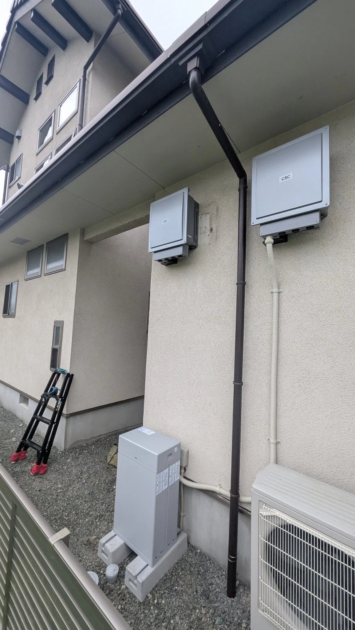 住宅の外壁に取り付けたパワーコンディショナ2台と屋外の切替盤。脚立を立てた施工中の様子