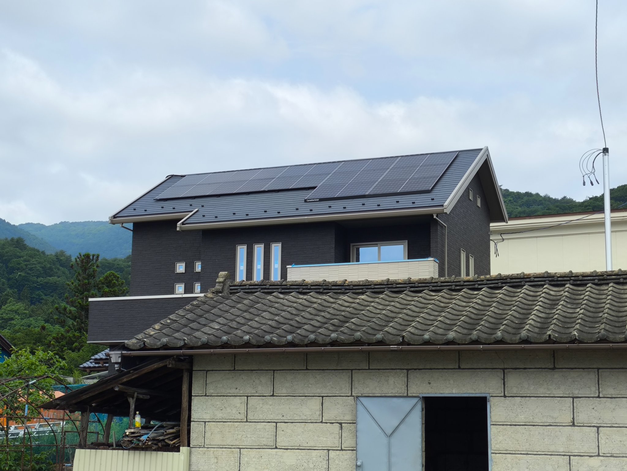 屋根に太陽光パネルを設置した戸建住宅の外観。手前に瓦屋根の納屋、背景に山